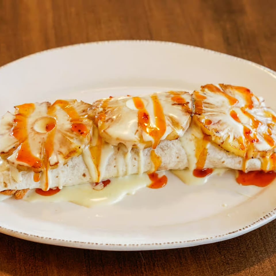 Burrito topped with grilled pineapple slices, melted cheese, and a drizzle of orange sauce on a white plate.