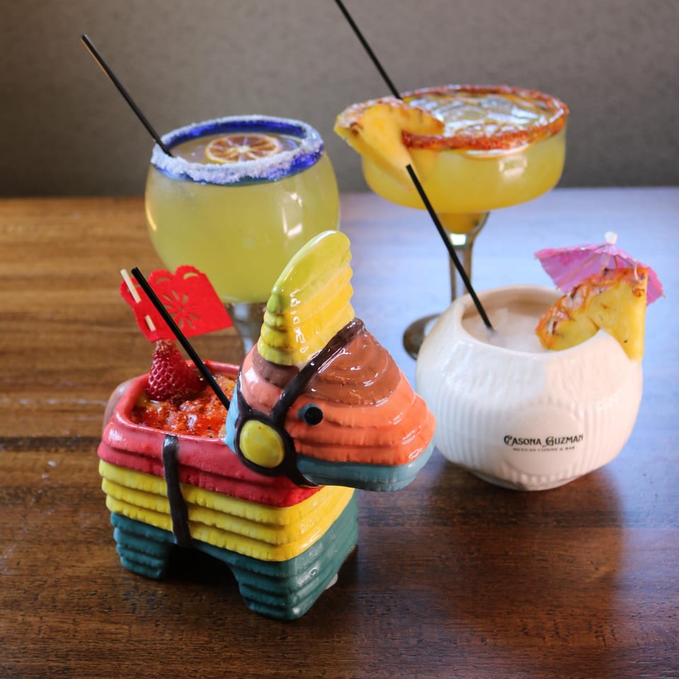 Three colorful cocktails, including one in a piñata-shaped glass, on a wooden table.