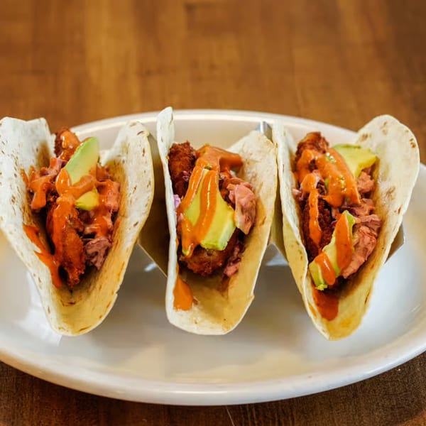 Three tacos with fried chicken, pulled pork, avocado, and orange sauce on a white plate.