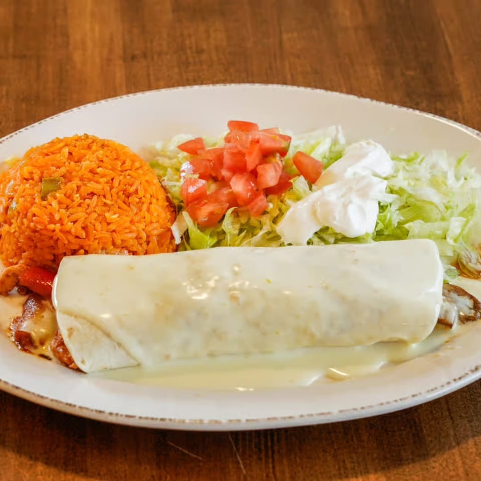 A large burrito covered in white cheese sauce, with a side of orange rice, lettuce, tomatoes, and sour cream.